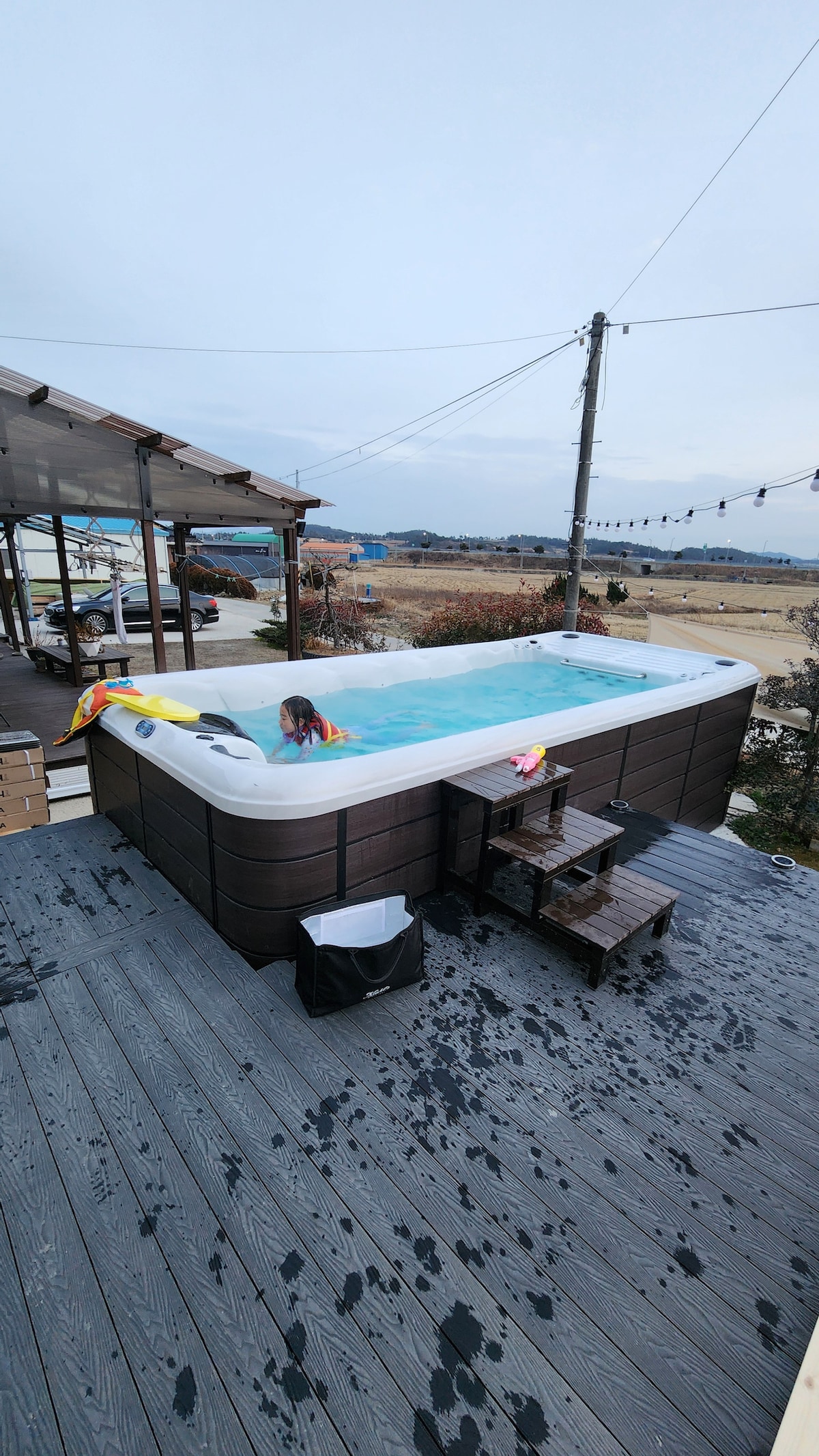 A hot tub is situated on a wooden deck, surrounded by an open landscape. Steps lead up to the hot tub, which is partially filled with water. Colorful inflatable pool toys are visible on the edge, enhancing the outdoor relaxation space.
