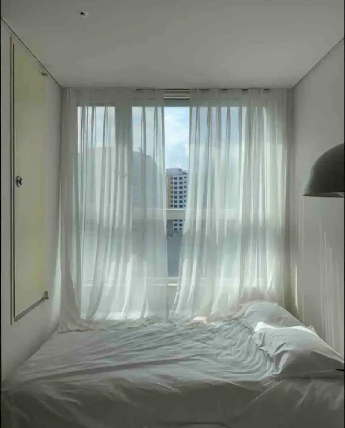 A bright sleeping area features a spacious bed dressed in light linens. Sheer white curtains frame a large window, allowing natural light to filter into the room. Soft shadows are cast on the walls, creating a calm and soothing environment.