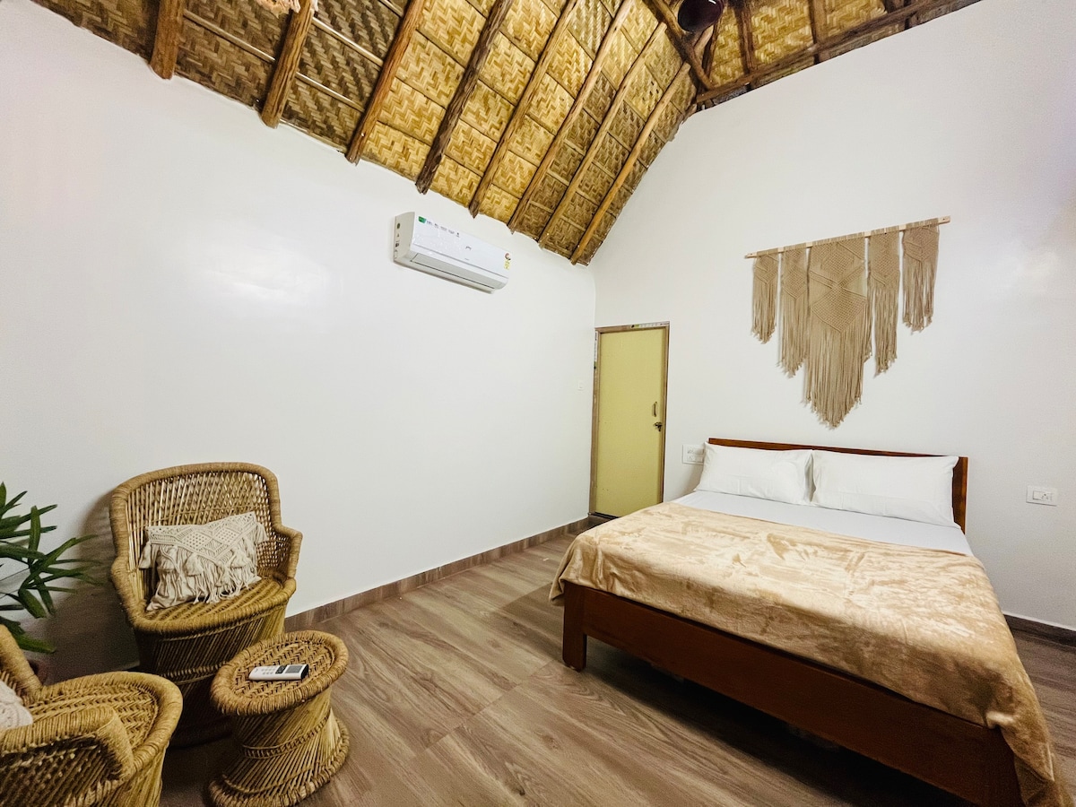 A cozy bedroom features a spacious bed dressed in soft linens, set against a plain wall. Two woven chairs are positioned nearby, complemented by a decorative macrame wall hanging. A ceiling with a natural thatched roof adds warmth to the ambiance, while an air conditioning unit is mounted above.