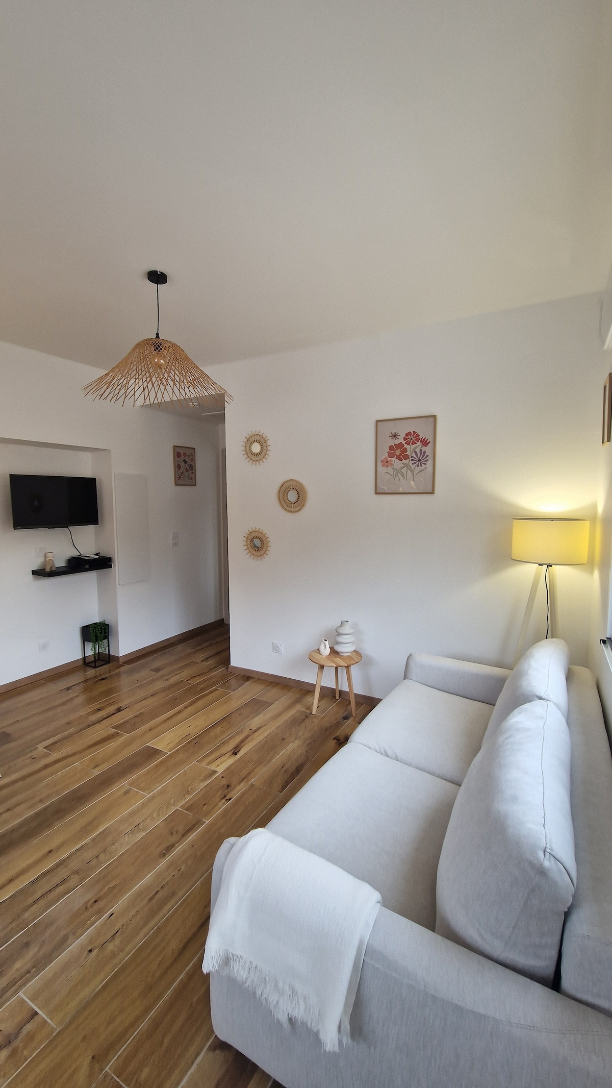 A cozy living area features a light gray sofa complemented by a small wooden side table. A charming lamp provides soft illumination, while art adorns the walls. Natural wood flooring adds warmth, and a wall-mounted TV is visible to one side.
