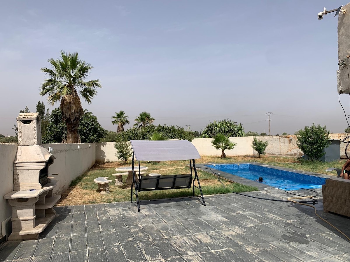 An outdoor area features a swimming pool surrounded by grass and several palm trees. A stone barbecue stands nearby, alongside a set of circular tables and chairs. A shaded swing seat adds to the relaxation space, while a gentle breeze can be felt in this tranquil setting.
