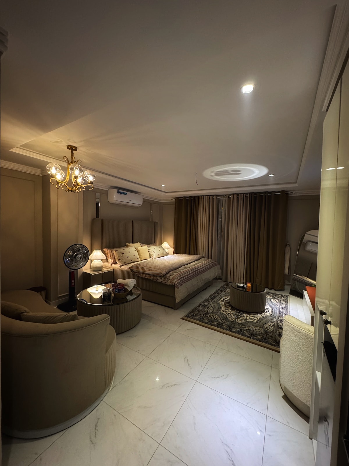 The studio features a spacious bed with decorative pillows and a patterned throw blanket. Two comfortable armchairs are positioned near a round side table. Soft lighting is provided by a chandelier and lamps, while large curtains frame a window, enhancing the room's ambiance.