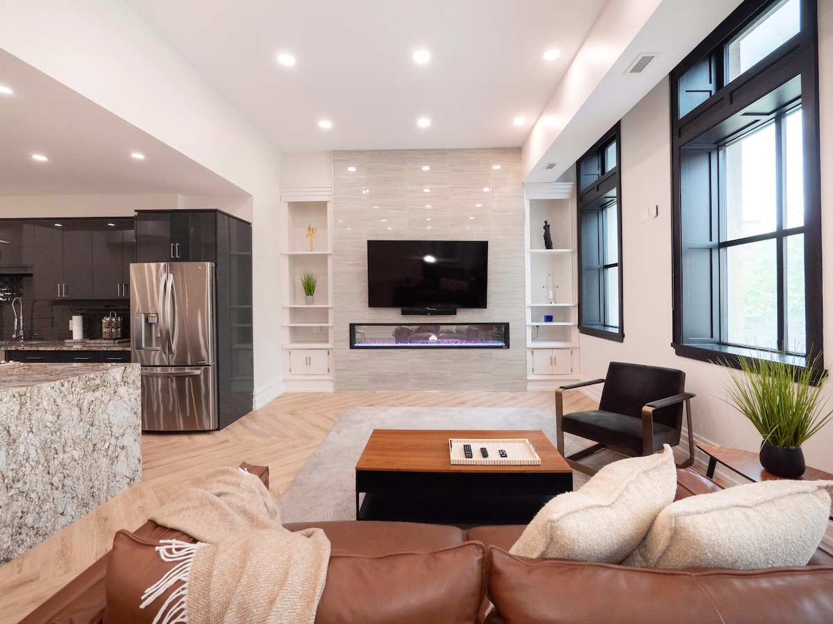 A spacious living area features large windows allowing natural light to flood in. A sleek electric fireplace is mounted on a light-colored stone wall, while a brown leather sofa is accented with soft throw pillows. A modern coffee table sits in the center, inviting relaxation.