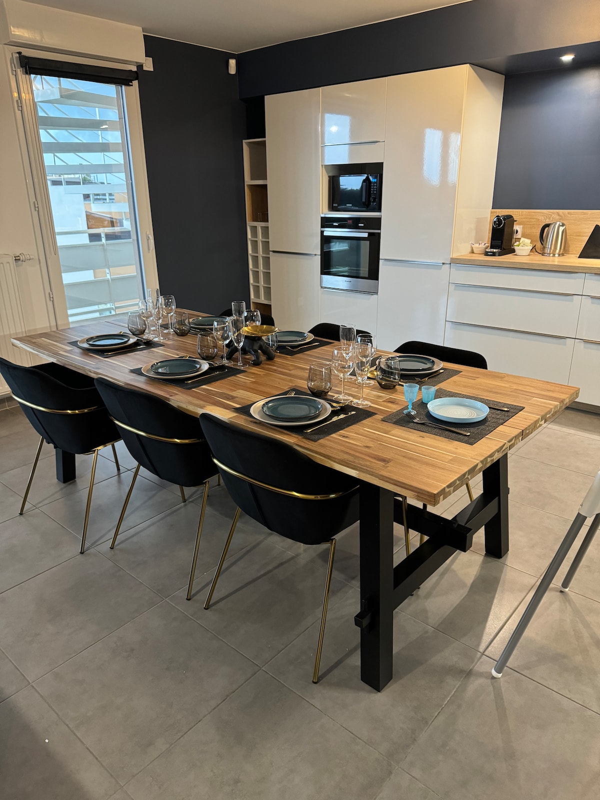 A dining area is presented with a wooden table, set for a meal with plates and glassware. Six black chairs with gold accents surround the table. The kitchen is visible in the background, featuring modern appliances and cabinetry.