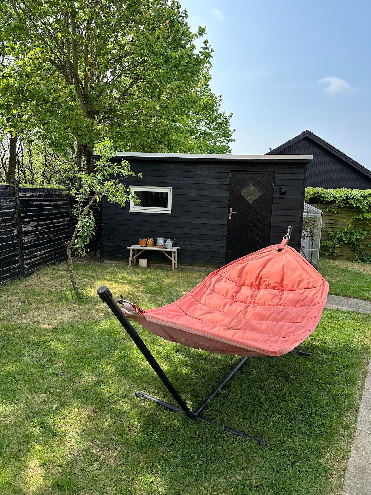 A serene outdoor area features a vibrant orange hammock chair positioned on lush green grass. A small outdoor table holds utensils, and a black shed is visible in the background, accented by a nearby tree and a wooden fence.