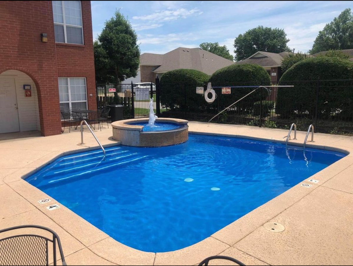 A saltwater pool surrounded by a tiled deck is visible, featuring a central fountain. A secured fence creates a boundary, with landscaped shrubs providing privacy. Several lounge chairs are positioned around the pool, allowing for relaxation in a tranquil outdoor setting.