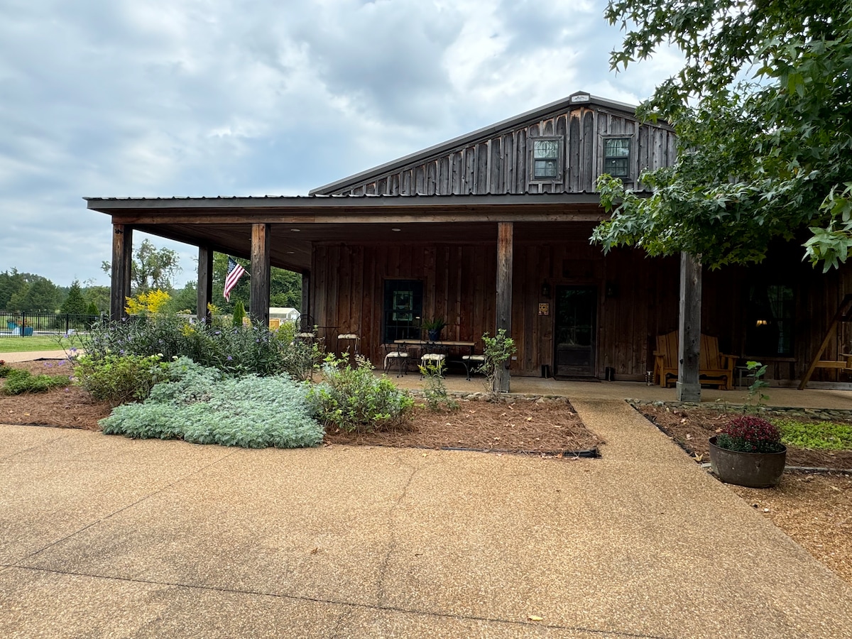 A rustic wooden home is set against a cloudy sky, featuring a wide porch with seating. Lush greenery and flower beds border the entrance, while a pathway made of gravel leads to the front door, inviting guests to explore the outdoor space.