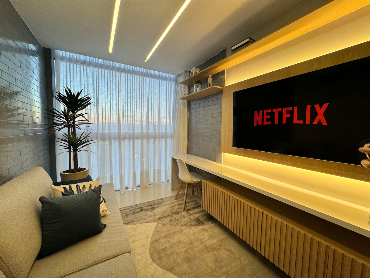 A cozy living area features a light-colored sofa adorned with decorative cushions, positioned near a sleek entertainment unit displaying a Netflix logo. Natural light filters through sheer curtains, illuminating the warm tones of the wooden shelving and the subtle texture of the area rug.