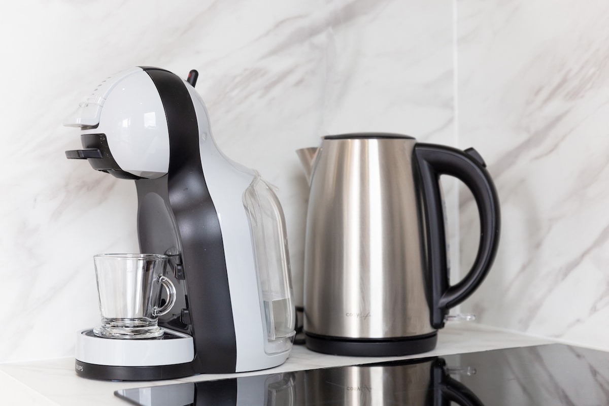 A modern coffee machine is positioned beside a sleek metallic kettle on a countertop. The coffee machine is designed with a clear water reservoir, and an empty glass sits in front of it, ready for use. The backdrop features glossy marble-effect tiles.