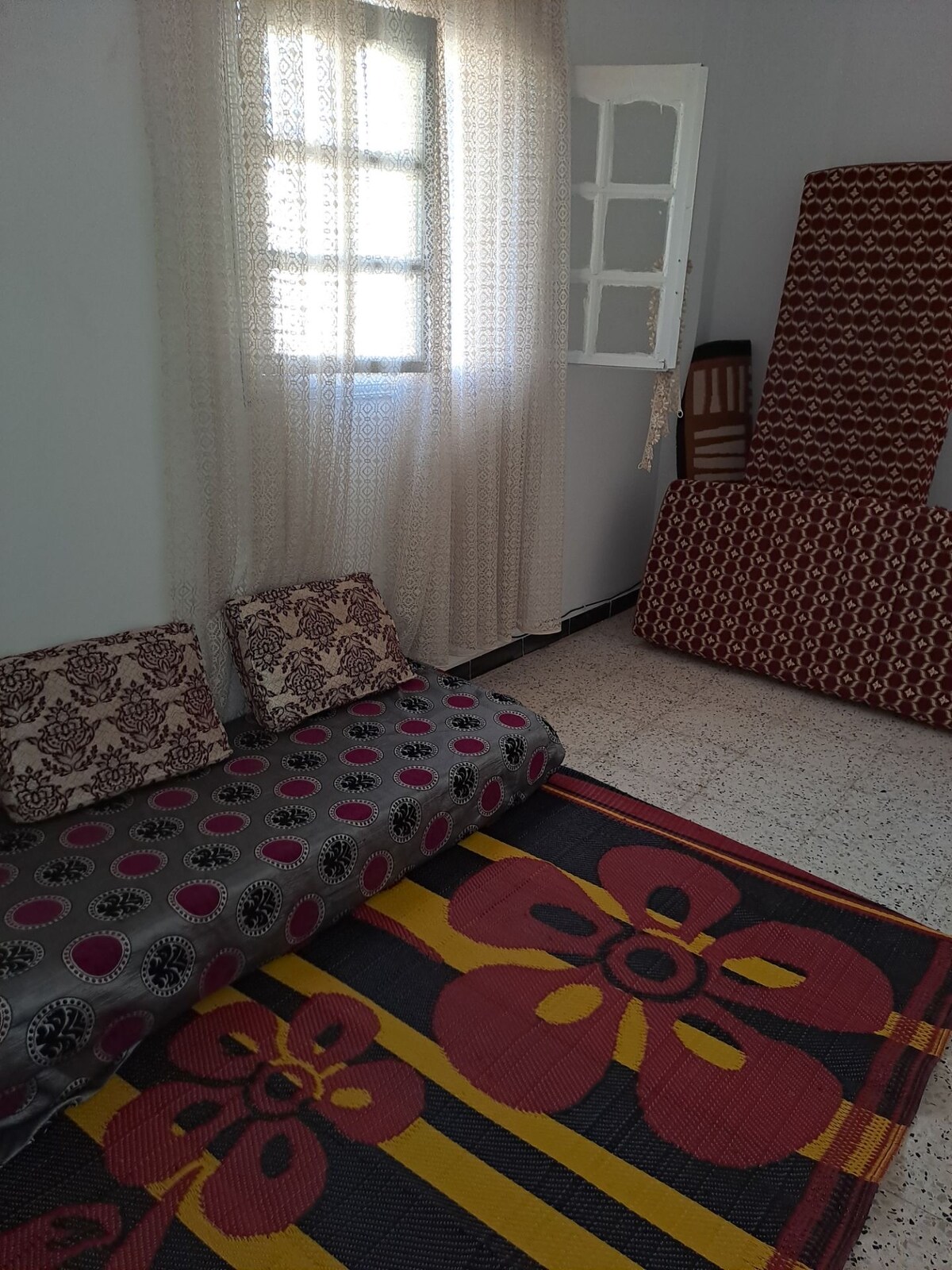 A simple seating area is presented with upholstered cushions placed against the wall. A colorful woven rug adorns the floor, displaying floral motifs. Light filters through sheer curtains, while a partially closed window adds a touch of natural light to the space.