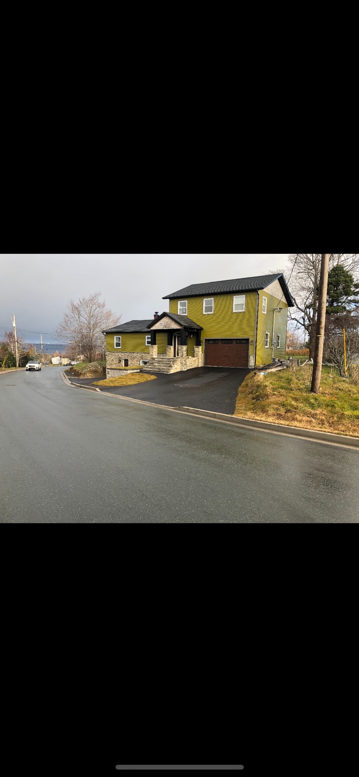 The exterior of a two-story house is visible, painted in a distinctive green color. A sloped driveway leads to a garage, while the surrounding area features a mix of grass and pavement. The house is situated on a curve of the road, framed by trees.