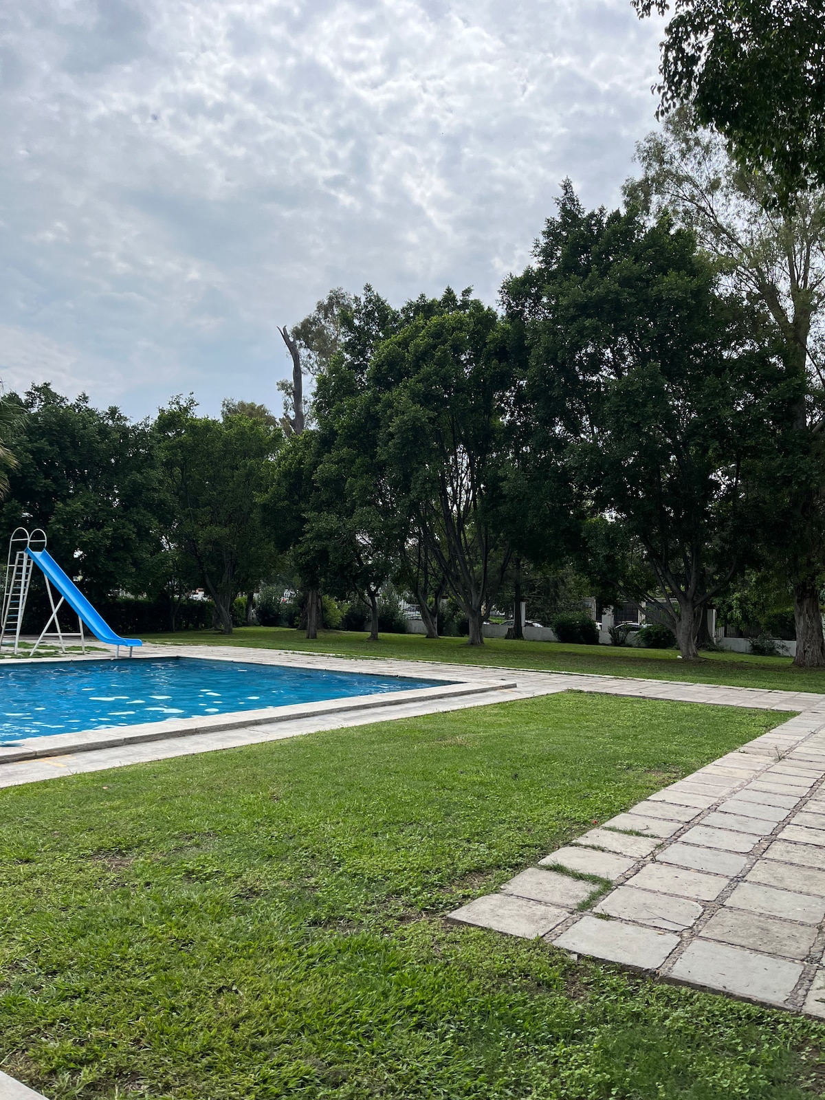 An outdoor swimming pool is visible, complemented by a blue water slide. Surrounding the pool, grassy areas are bordered by a stone patio, while trees provide shade in the background under a cloudy sky.