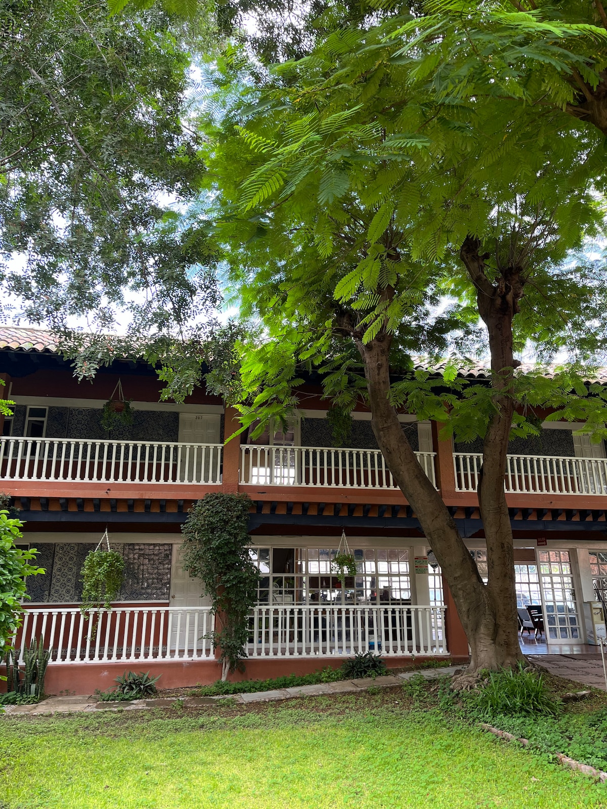 The exterior of a charming multi-level building is surrounded by lush greenery and tall trees. Balconies with white railings provide a view of the garden below. Soft sunlight filters through the leaves, creating a serene outdoor atmosphere.
