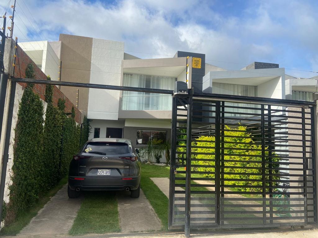 A modern home is presented behind a gated entrance, featuring a neatly manicured lawn and vibrant greenery. A parked car is visible in the driveway, while large windows allow natural light to fill the interior spaces.