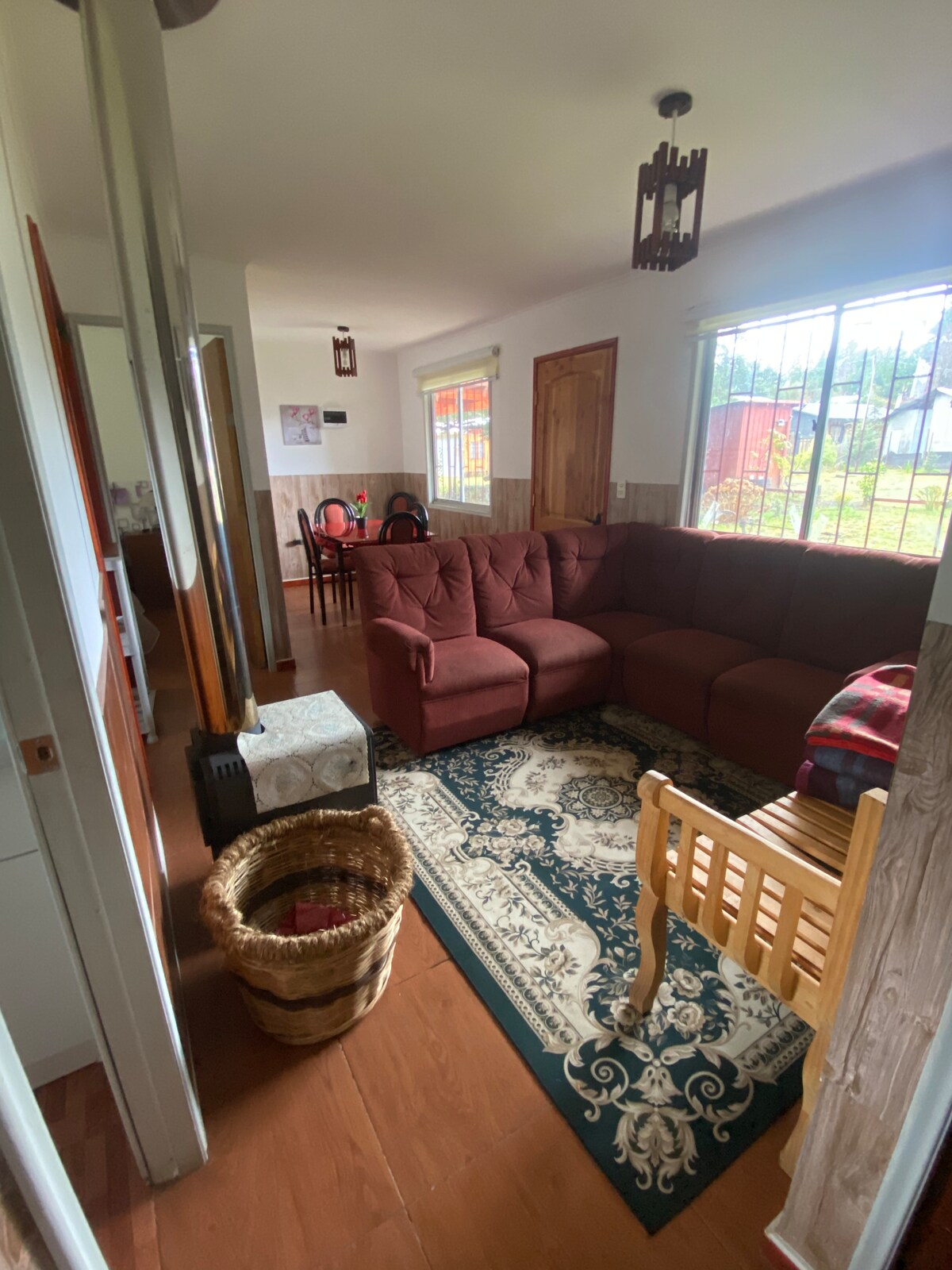 A cozy living area is organized with a large, L-shaped sectional sofa in a rich burgundy hue. A patterned area rug lays beneath, complementing a wooden table with legs visible. Natural light fills the space through large windows, enhancing the simple decor.