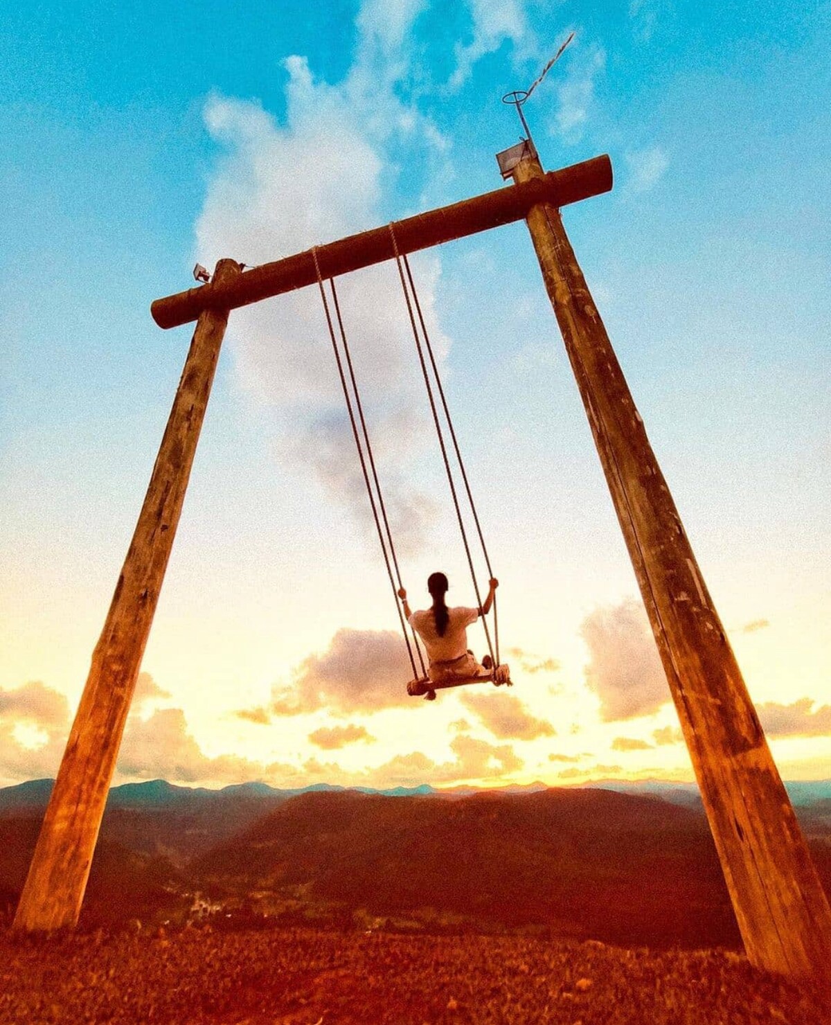 A wooden swing is positioned against a backdrop of a sunset, casting a warm glow over the surrounding landscape. The swing is framed by tall posts, emphasizing the openness of the scenery, with mountains in the distance under a colorful sky.