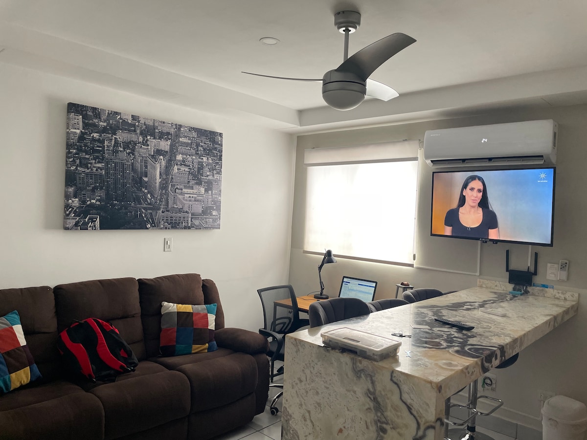 A modern living space features a brown sectional sofa adorned with colorful pillows. A sleek, marble-topped bar separates the living area from a small desk and chairs, with an air conditioning unit and a wall-mounted TV visible. Natural light filters through the window.