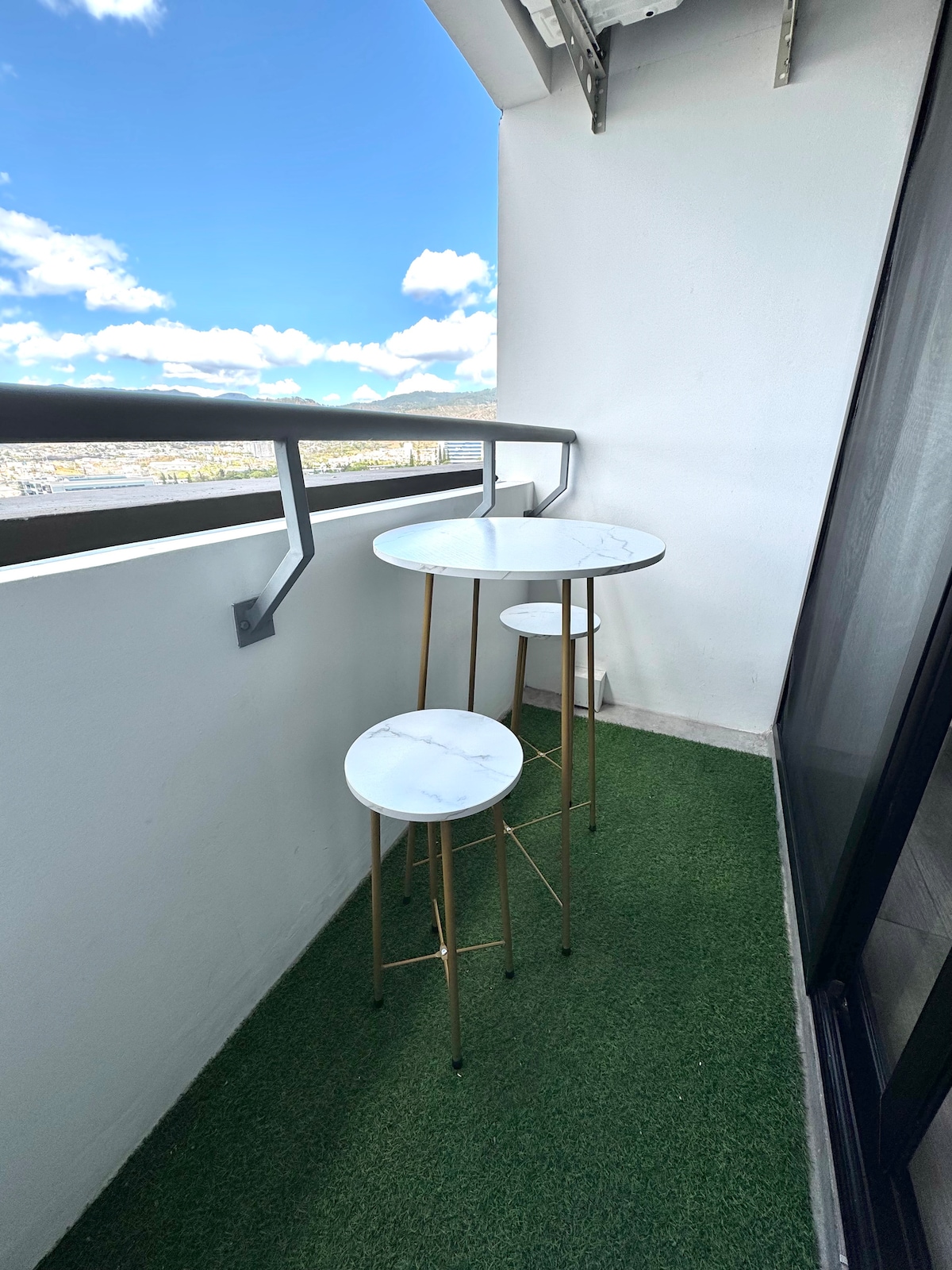A small balcony is presented with a green grass-like flooring and two marble-top tables with gold-colored legs. A clear sky dotted with white clouds is visible beyond the railing, creating a scenic outdoor space.