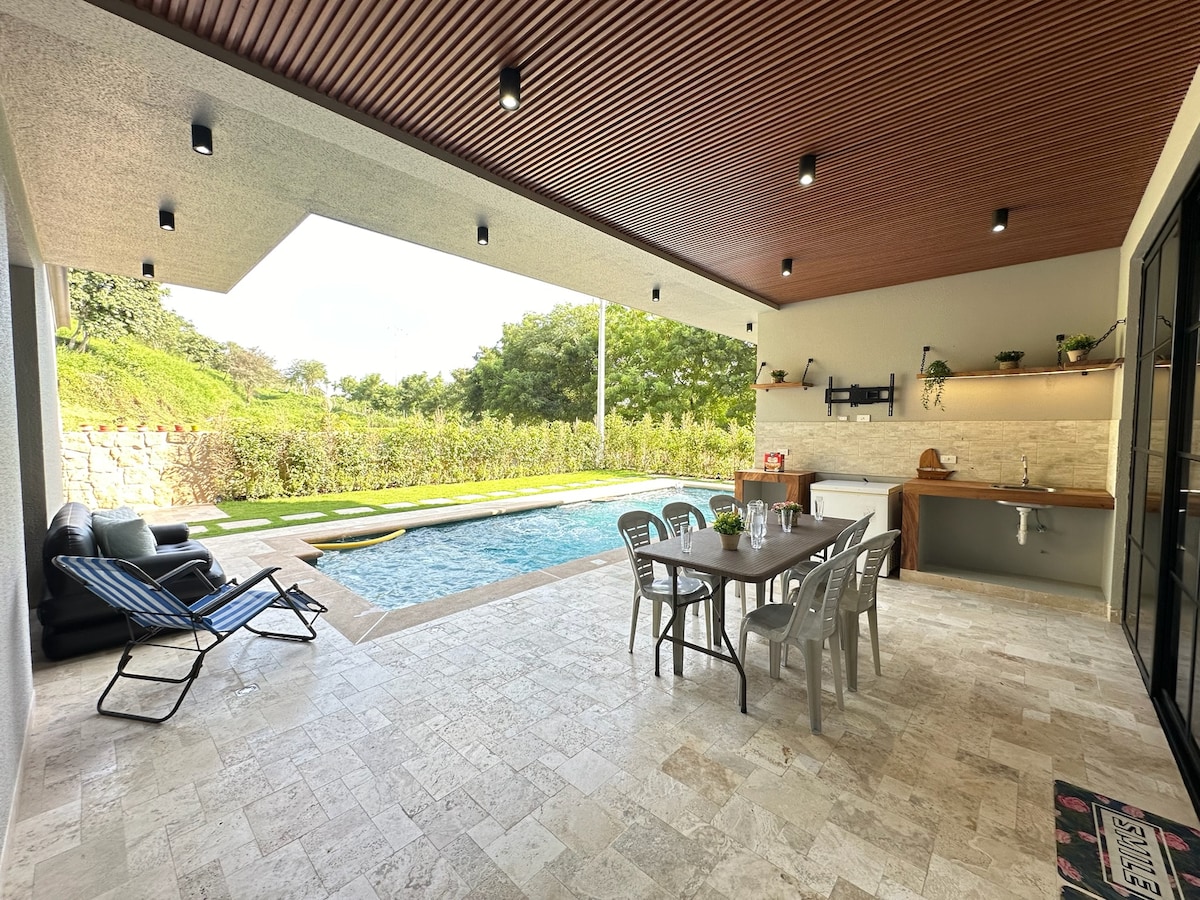 An outdoor patio area features a spacious dining table surrounded by metal chairs, ideal for meals al fresco. A refreshing pool is visible in the background, complemented by lush greenery. Comfortable lounge chairs are positioned nearby, offering a place to relax and enjoy the surroundings.