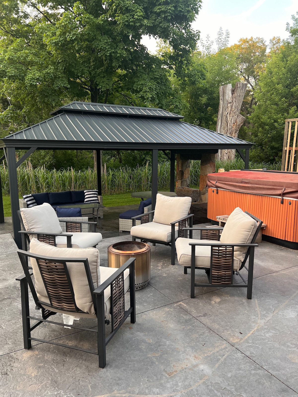 An outdoor seating area features four armchairs arranged around a firepit. A gazebo provides shelter, while a hot tub is positioned nearby. Lush greenery surrounds the space, creating a private, relaxing atmosphere for gatherings.