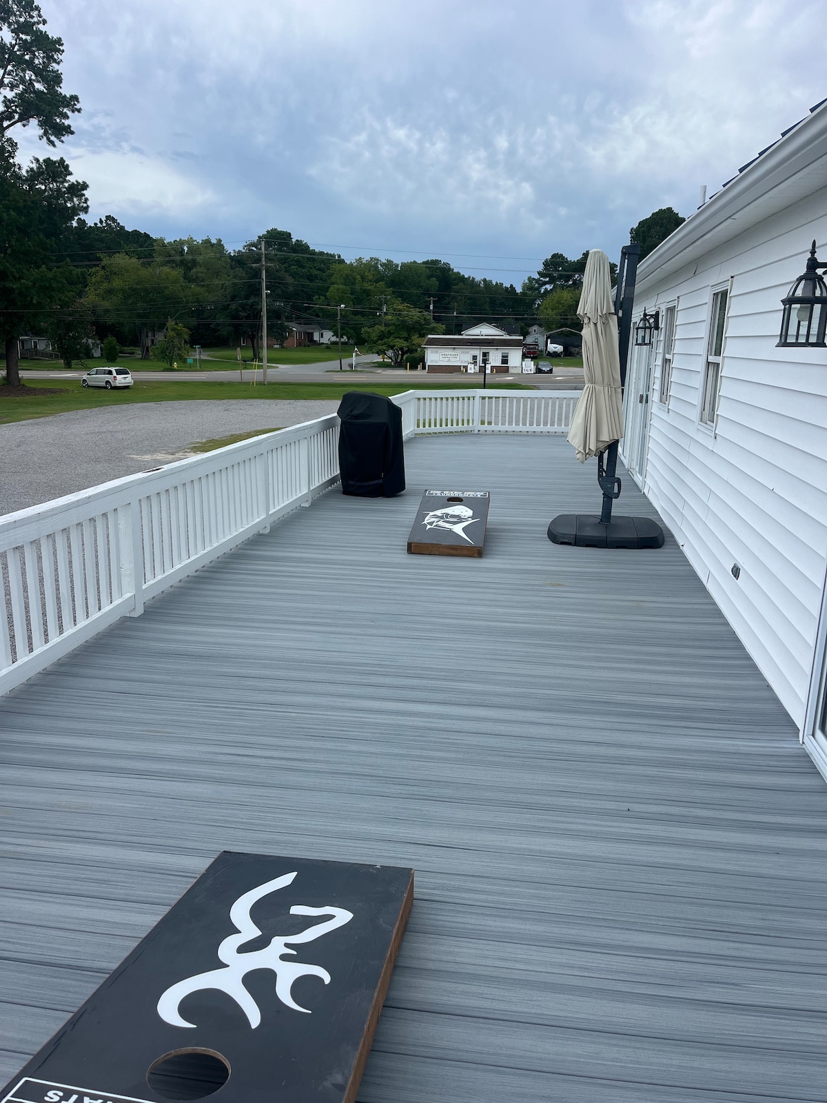 A spacious deck is depicted, featuring two cornhole boards positioned on a gray composite surface. A patio umbrella is placed in a base, providing shade. A barbecue grill is visible, and green trees are seen beyond the railing.
