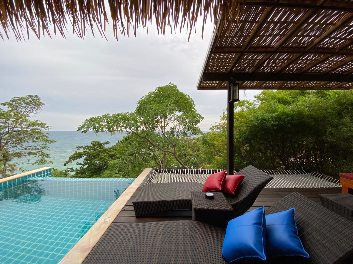 A spacious private pool area features comfortable lounge chairs, accented with red and blue cushions. The serene pool reflects the surrounding greenery, with a view of the ocean in the distance, framed by lush trees and a covered outdoor space.