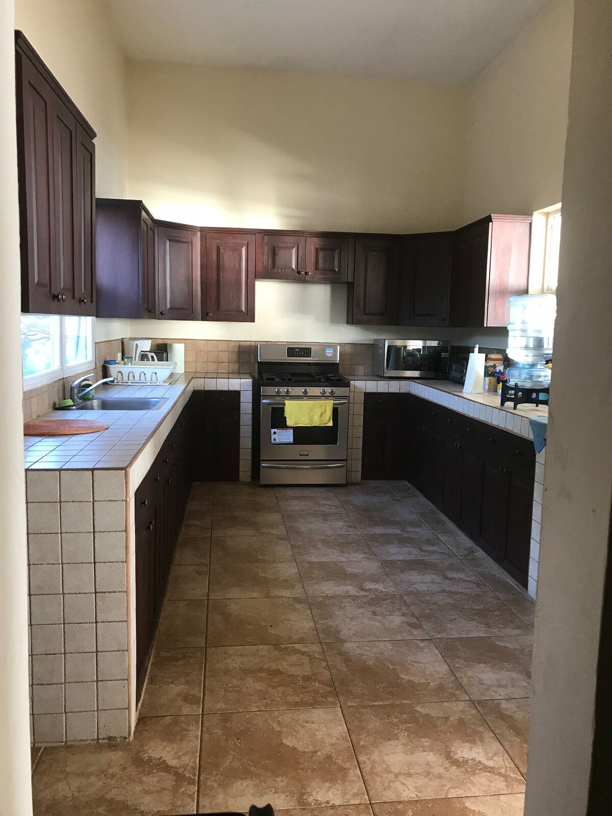 A spacious kitchen features dark wooden cabinetry and a tiled countertop. An oven and stovetop are positioned centrally, with a microwave and sink along the side. Light enters through a window, illuminating the tiled floor and creating a bright cooking space.
