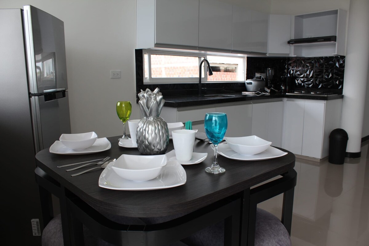 A modern kitchen is featured, showcasing a sleek black countertop and contemporary cabinetry. A dining table is set with white plates, colorful glasses, and utensils. The kitchen area includes stainless steel appliances, while large windows allow natural light to brighten the space.