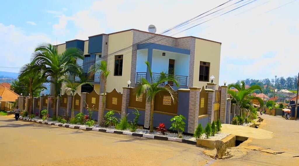 A modern multi-story building is situated on a wide street, surrounded by well-maintained landscaping. Palm trees and flowering plants are featured along the property line. The structure showcases a mix of pastel colors and large windows, creating a welcoming appearance.