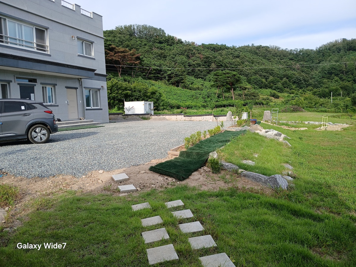 A spacious outdoor area is visible, featuring a gravel driveway next to a well-maintained lawn. A path made of stepping stones leads towards a landscaped yard with stones and grass, while a lush green hillside serves as a backdrop.