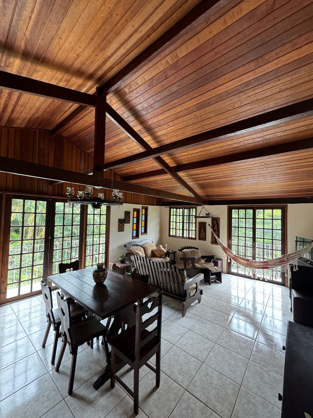 An open-plan living area is showcased, featuring wooden beams and a high ceiling. Large windows allow natural light to fill the space, while a dining table and seating area are arranged for comfort. A hammock is subtly included for relaxation.