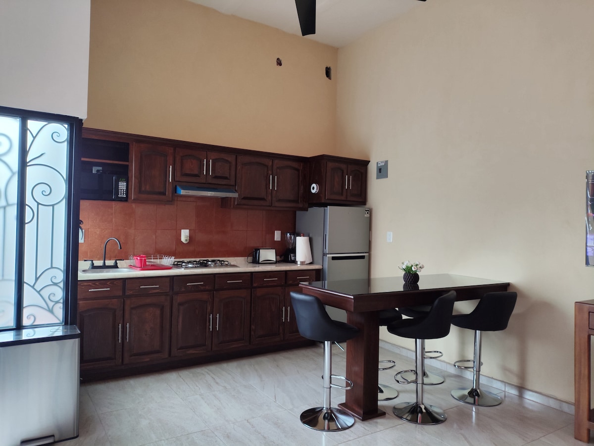 A kitchen space is complemented by dark wooden cabinetry and modern appliances. A dining table with black chairs is positioned adjacent to the kitchen, creating a functional area for meals. Natural light enhances the open layout, highlighting the marble flooring.