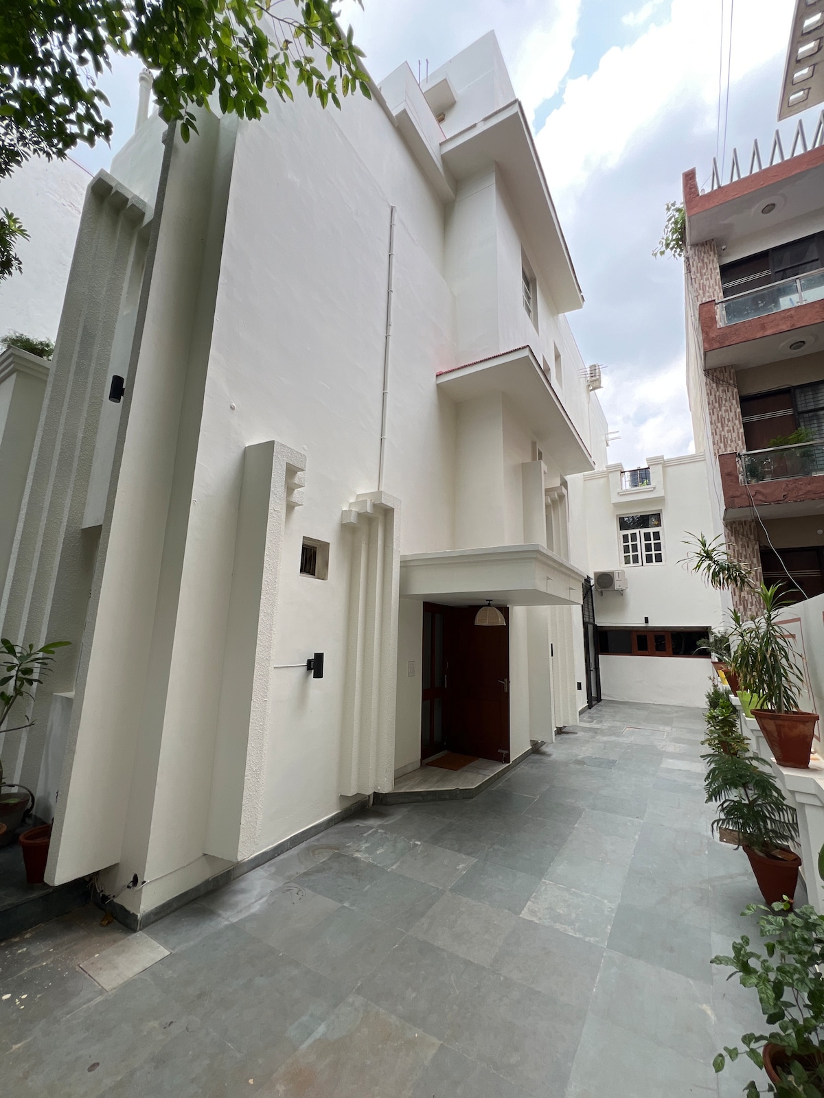 The exterior features a modern design with clean lines and a smooth white façade. A pathway made of stone tiles leads towards the entrance, flanked by potted plants that enhance the inviting atmosphere. Natural light filters through the surrounding greenery.