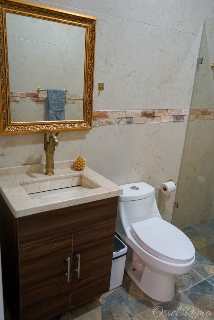 A modern bathroom is showcased with a wooden vanity featuring a stone sink. A vintage-style gold-framed mirror hangs above the sink. Adjacent, a white toilet is positioned next to a small waste bin. The tiled floor adds a clean and polished look to the space.