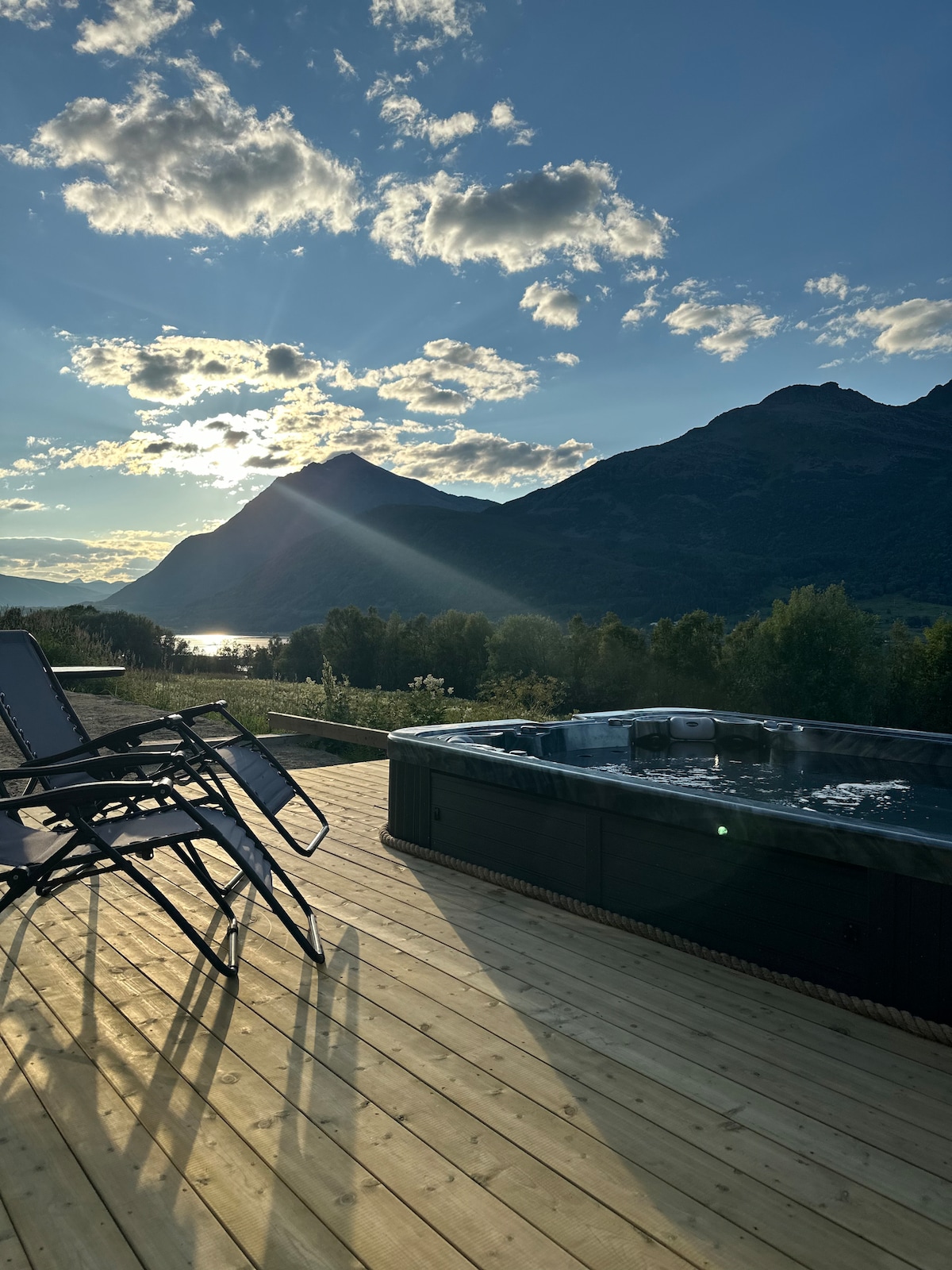 A wooden deck features a hot tub with surrounding mountains and a clear sky in the background. Two lounge chairs are positioned nearby, allowing for relaxing views of the natural landscape and the shimmering body of water below.