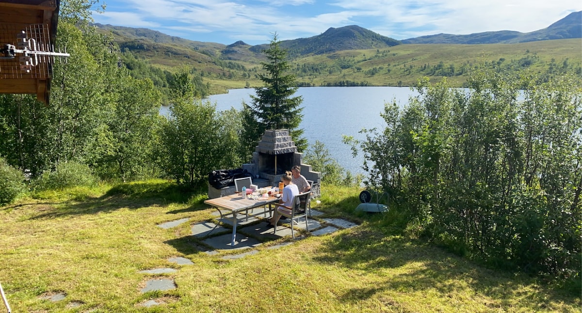 An outdoor dining area is set with a table and chairs, surrounded by greenery. A view of a serene lake and distant mountains is visible in the background under a clear blue sky. A barbecue grill is positioned nearby.