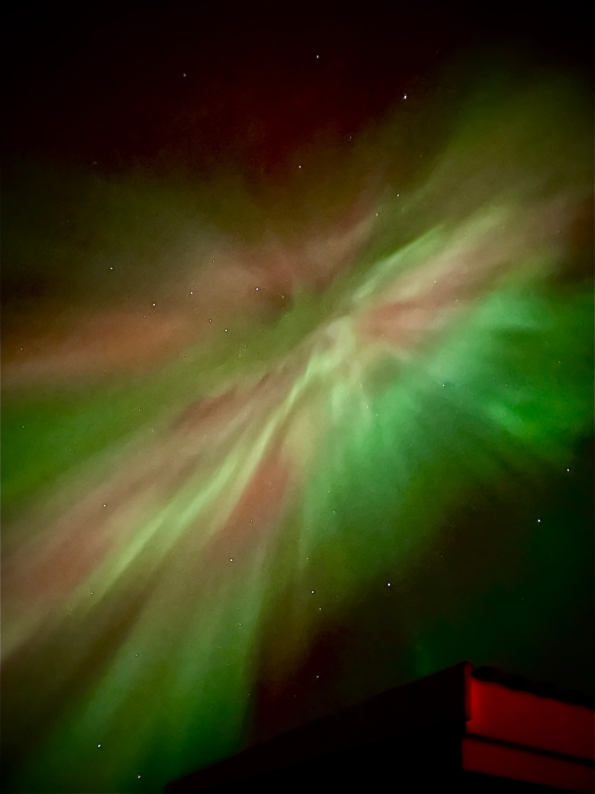 A vibrant display of the northern lights fills the night sky, showcasing swirling shades of green and hints of red against a backdrop of stars. The top edge of a building is visible at the bottom of the image.