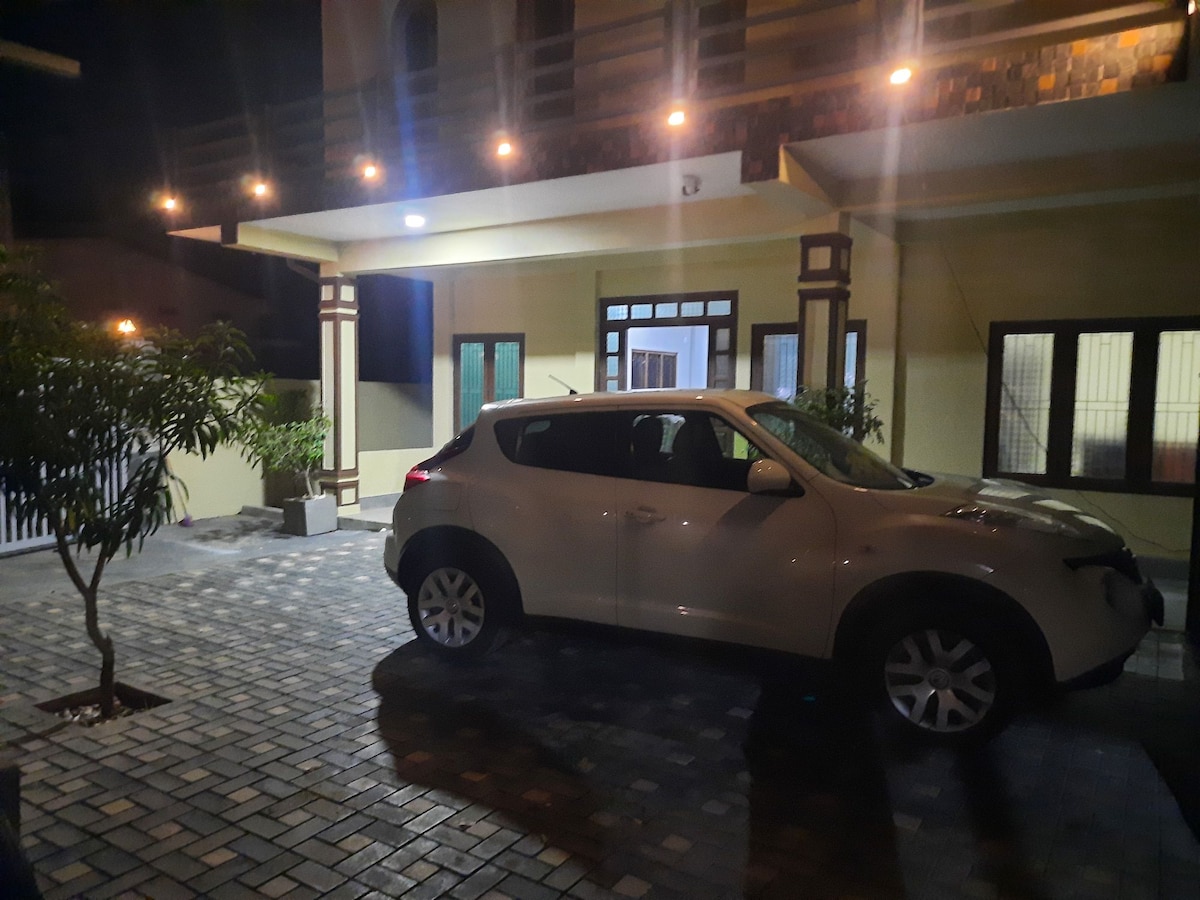 A well-lit driveway is depicted at night, showcasing a white vehicle parked under the home's covered entrance. Decorative outdoor lighting illuminates the surrounding area, highlighting the textured pavement and a small tree positioned nearby.