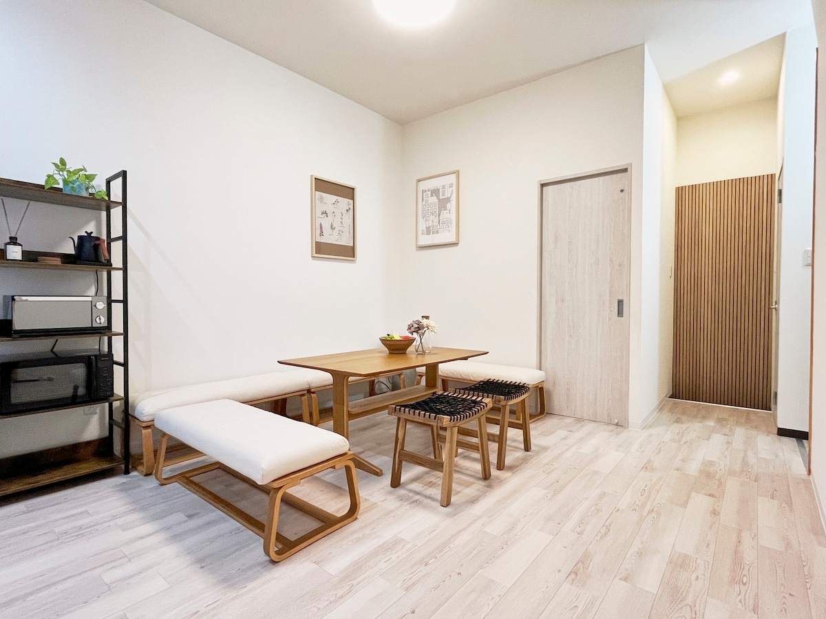 A well-lit dining area is furnished with a wooden table and comfortable benches. Minimalist decor elements, including framed art, adorn the walls. A doorway leads to another room, while a plant adds a touch of greenery to the space.