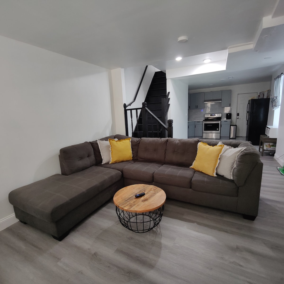 A modern living area features a spacious sectional sofa with yellow accent pillows, arranged around a circular wooden coffee table. In the background, a staircase leads to upper levels and a well-equipped kitchen is visible, showcasing sleek appliances and light cabinetry.