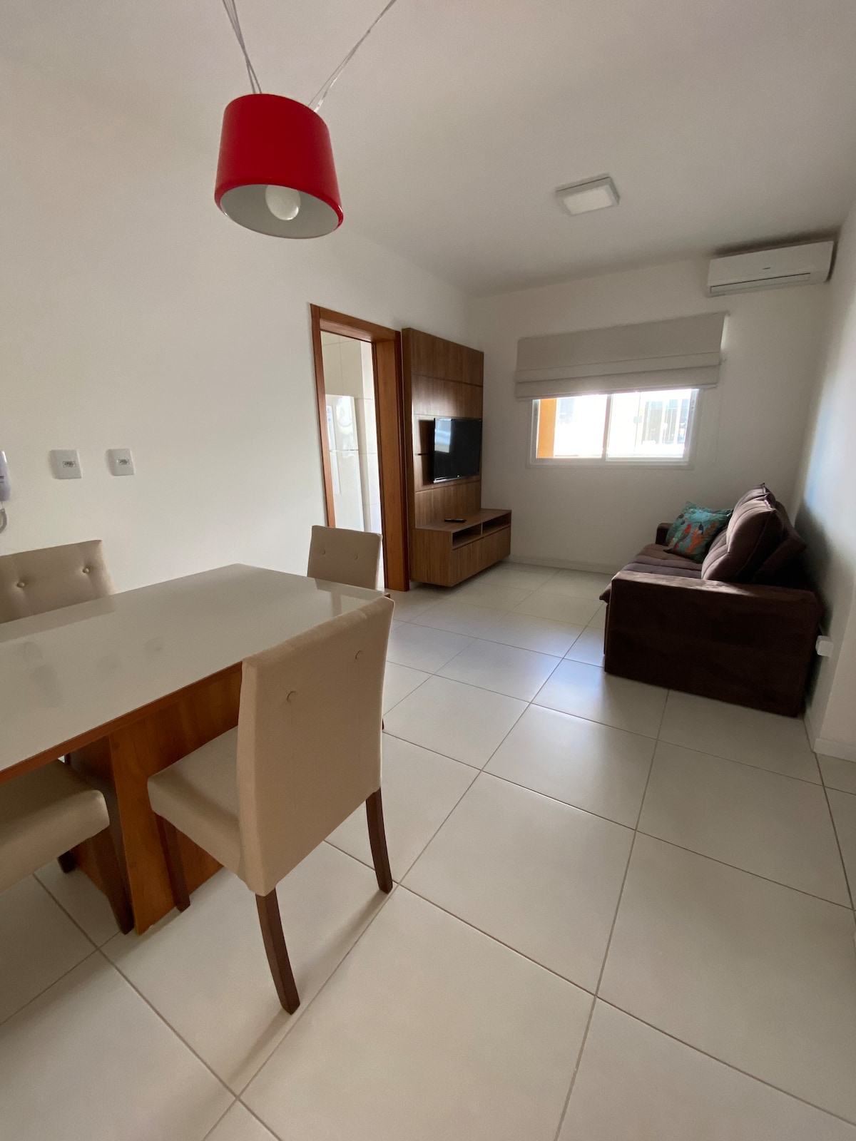 A living area features a compact brown sofa beside a wooden media unit with a television. A dining table with four upholstered chairs occupies one corner, while a window allows natural light to fill the space, complemented by neutral-colored walls.