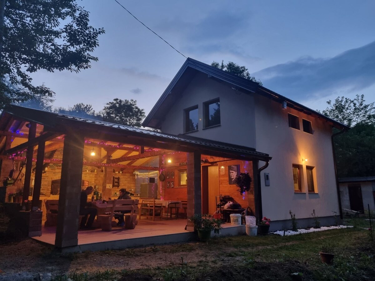 The exterior of the cottage is highlighted at dusk, featuring a spacious covered patio with wooden beams. Soft lighting enhances the atmosphere, while wooden tables and benches invite gatherings. Windows reflect the serene natural surroundings, surrounded by greenery and evening sky.