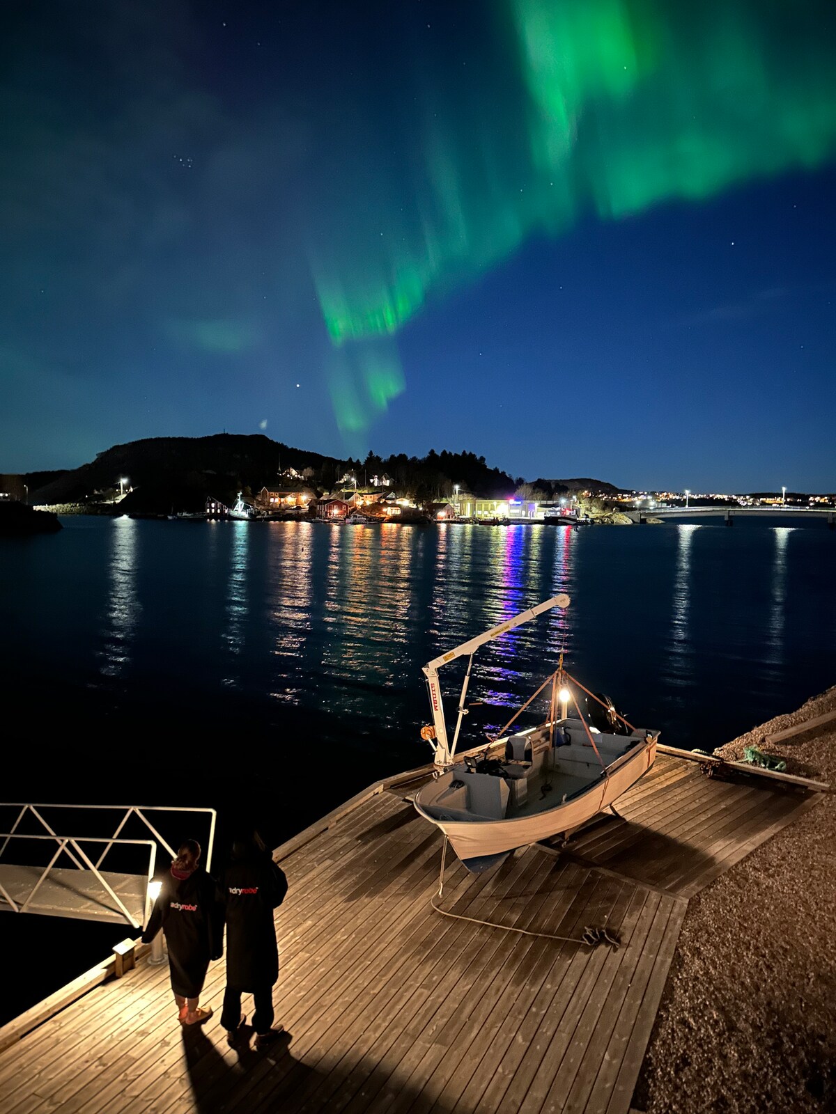 The night sky is illuminated by vibrant green auroras arching over a calm waterway. A small boat is anchored at a wooden dock, with distant lights from a nearby town reflecting on the water's surface. Two figures stand on the dock, gazing up at the display.