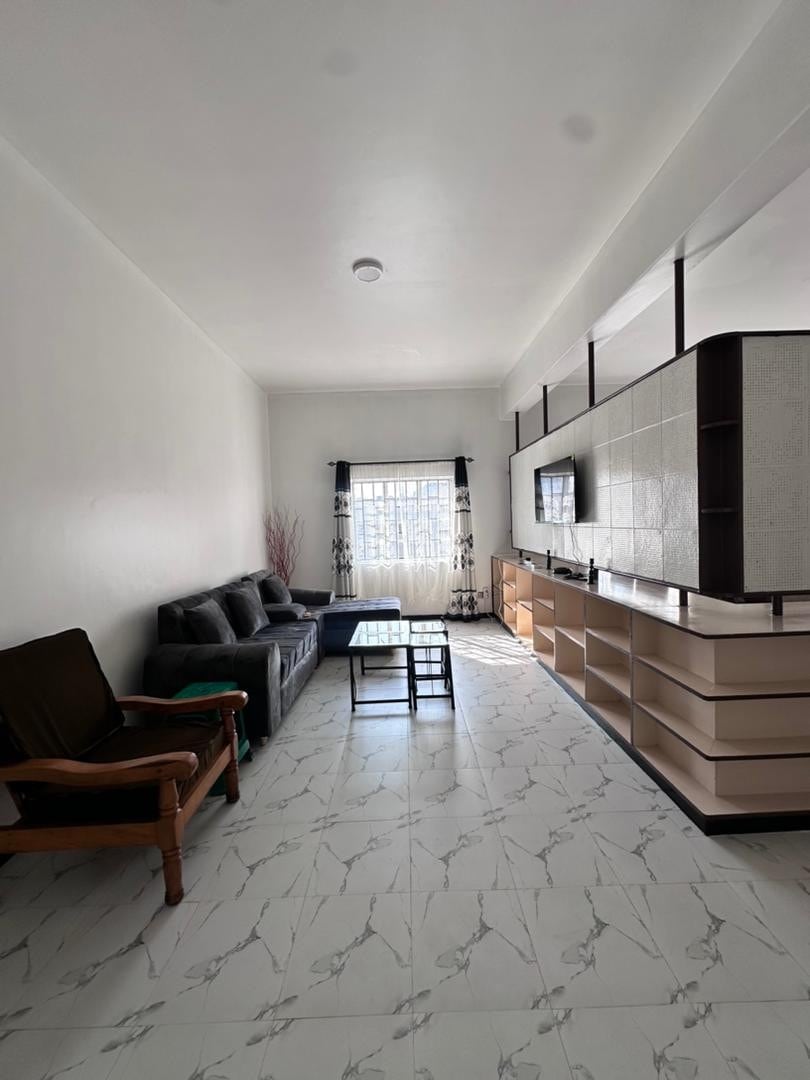 A living area features a dark sofa set and a small coffee table, with a media unit against the wall. Natural light enters through the window, which is framed by patterned curtains. The flooring is tiled with a marble-like design.