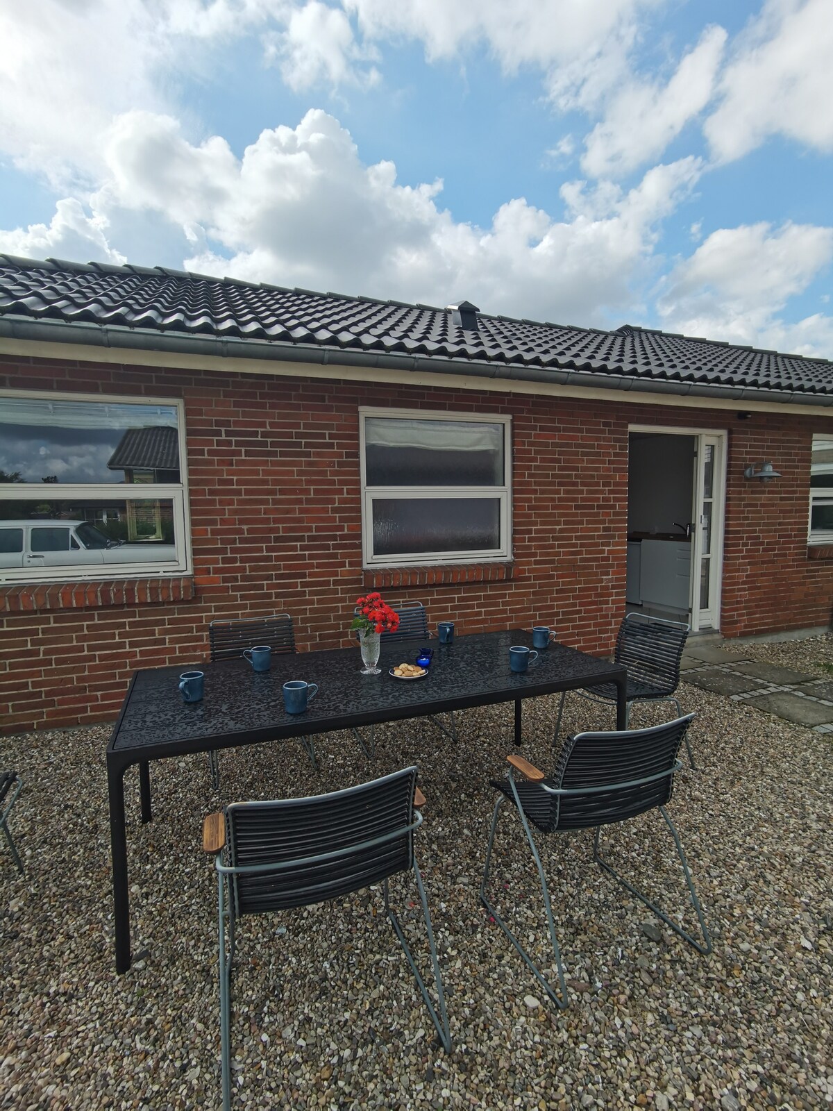 An outdoor dining area features a rectangular black table with several chairs arranged around it. A small vase with flowers sits on the table, accompanied by coffee cups. The background includes a brick wall and a sky filled with clouds.