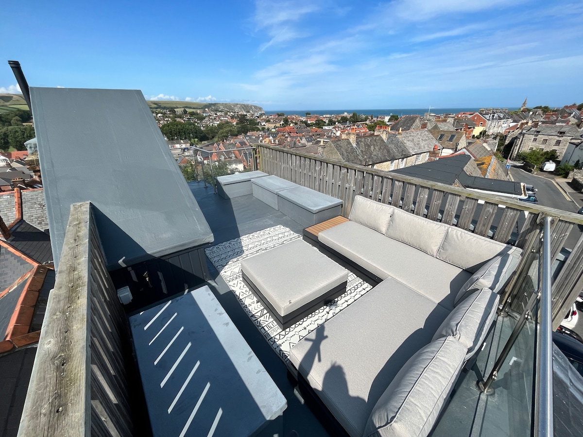The spacious roof terrace showcases comfortable outdoor seating and a central coffee table. Panoramic views of the surrounding area, including the coastline and rooftops, are visible under a bright blue sky, creating an inviting space for relaxation.