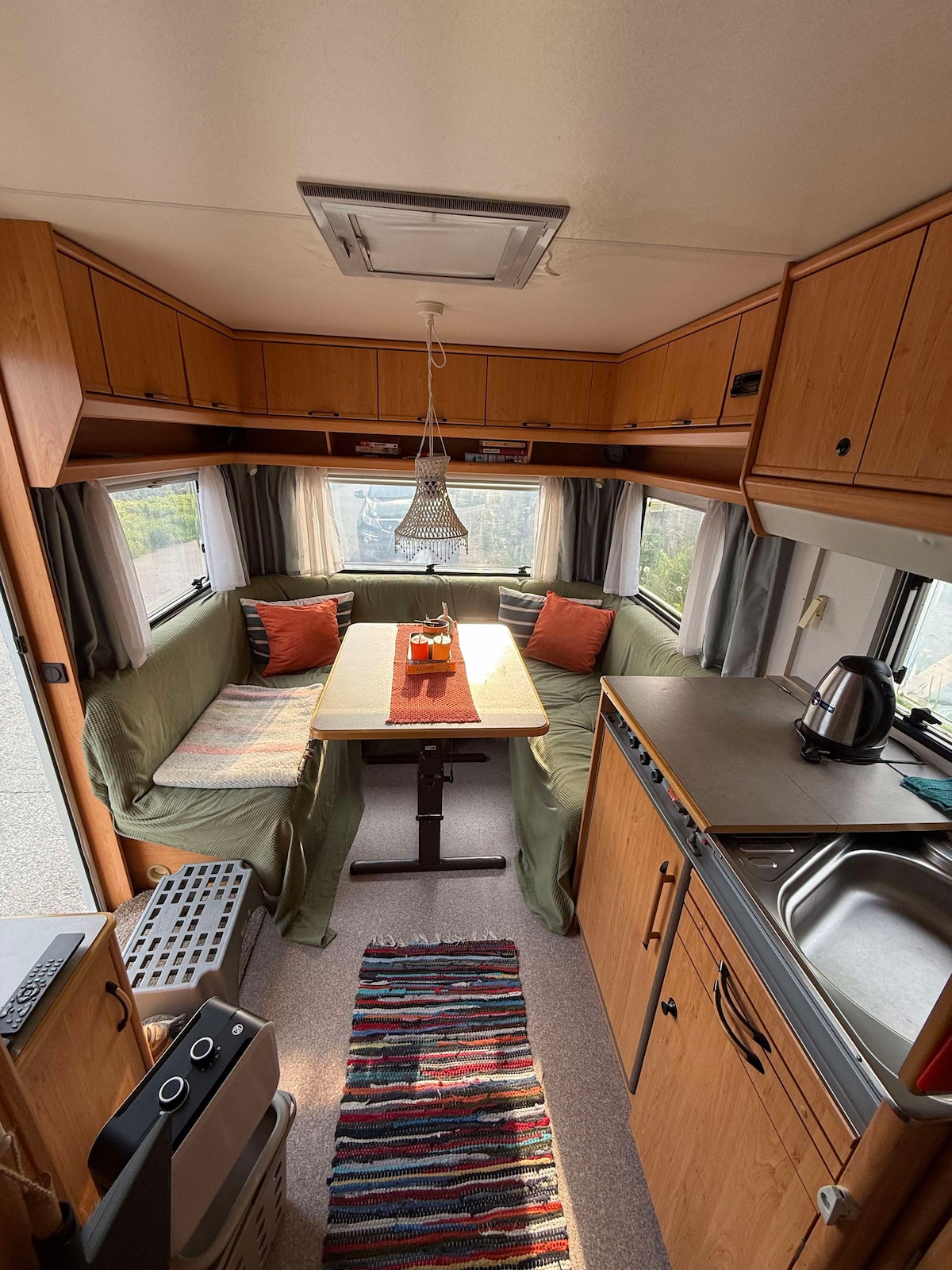 The interior of the camper is designed with wooden cabinetry and a cozy seating area, featuring a table surrounded by a long, upholstered bench and cushions. Natural light enters through window curtains, providing a view of the landscape outside. A small kitchenette is equipped with essential appliances.