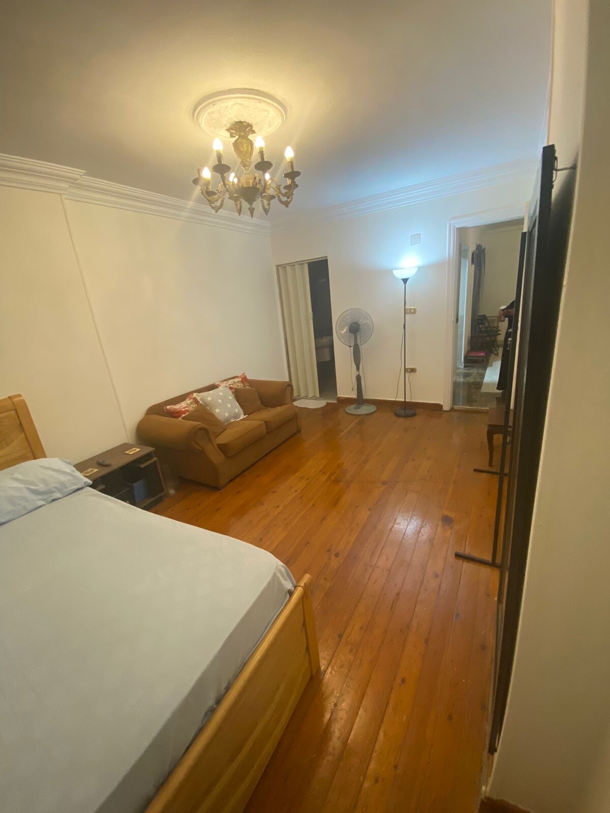 A small room features a hardwood floor and a spacious bed with light bedding. A decorative chandelier hangs from the ceiling, providing soft illumination. A brown couch is positioned near a standing fan, with a doorway leading to another space visible in the background.