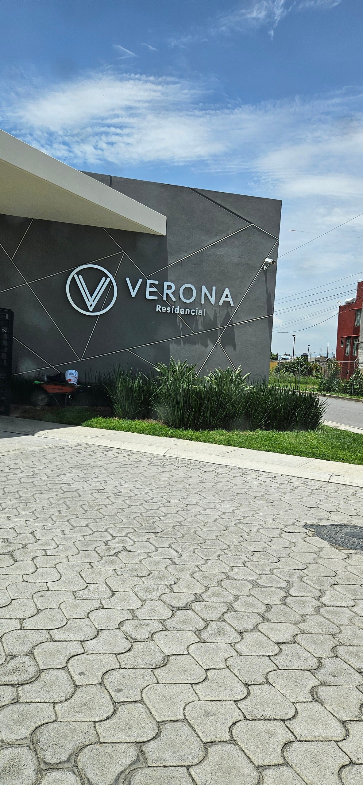 The exterior of a modern building features a prominent sign reading 'VERONA Residencial.' Paved walkways and manicured greenery create a welcoming entrance to the property under a clear blue sky.