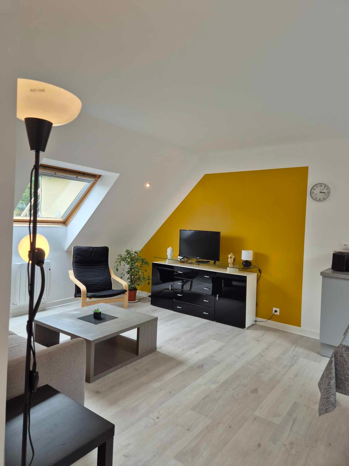 A bright living area features a sloped ceiling and a striking yellow accent wall. A modern black TV stand holds a flat-screen television, while a light wood coffee table sits at the center. A comfortable chair and a floor lamp add to the inviting decor.